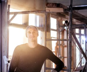 Attractive builder is standing next to scaffolding. He illuminated by sunbeams of sunset